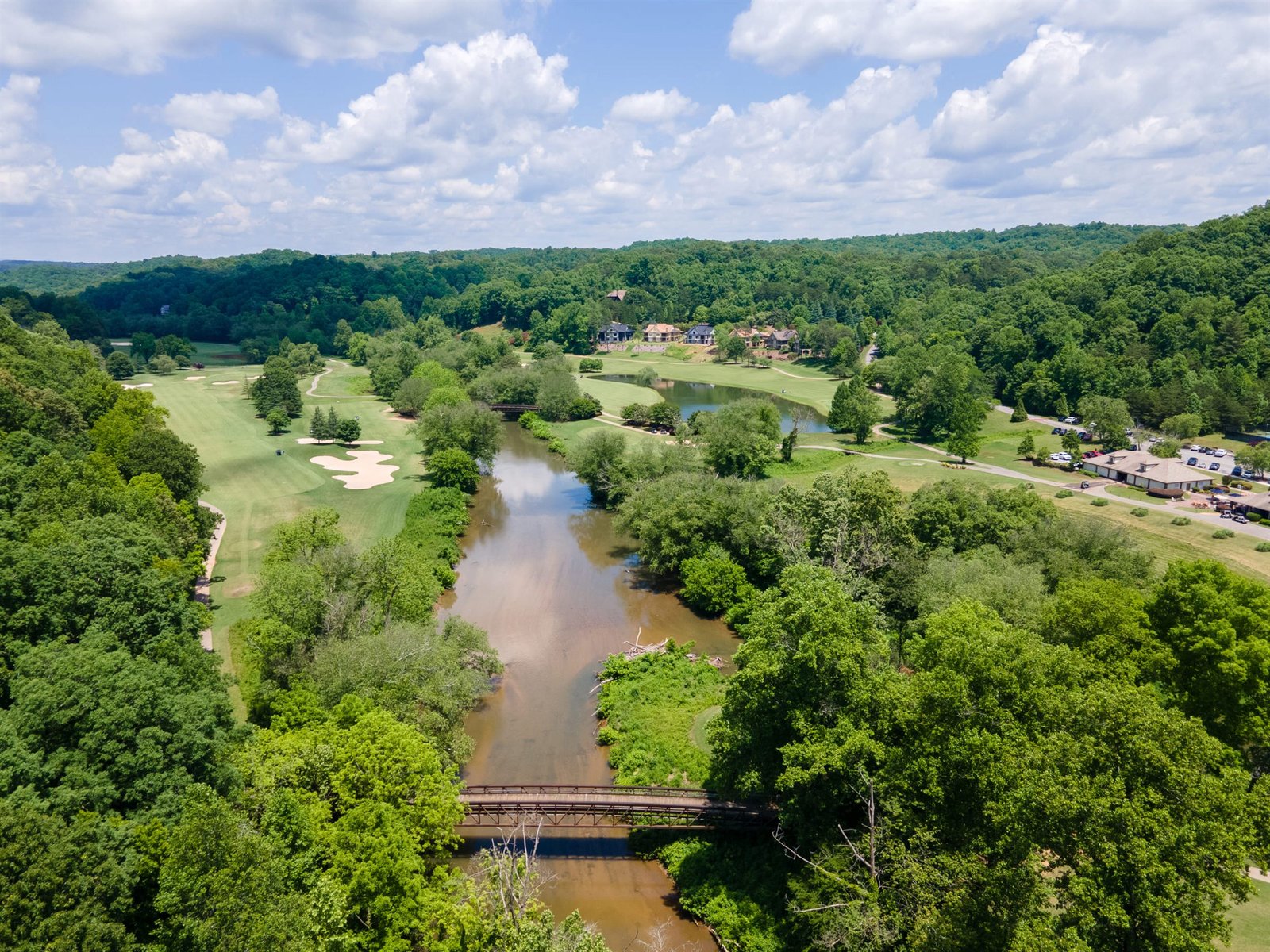 Aerial view of the Chestatee River winding through the Achasta golf course in Dahlonega Georgia &mdash; the design constraint that Nicklaus integrated into the back-nine routing, with a pedestrian footbridge crossing the water and estate homes nestled in the hardwood canopy along the bank