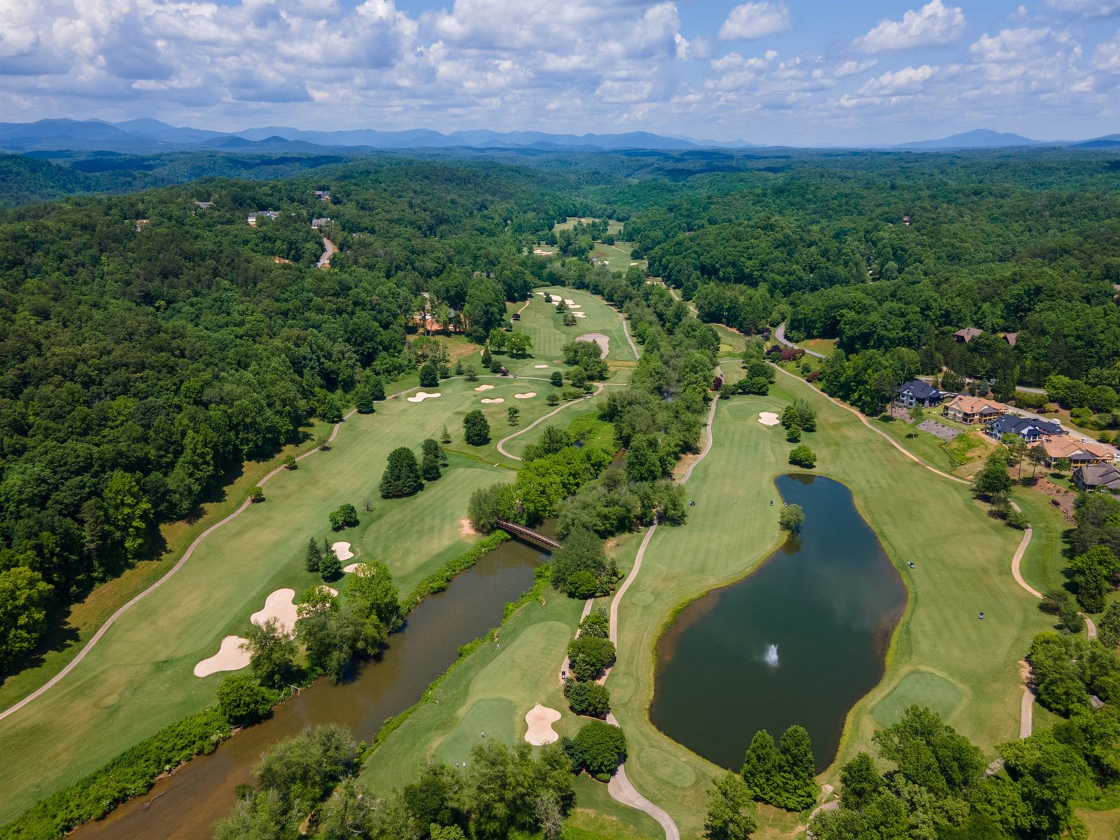 Aerial view of the Jack Nicklaus signature golf course at Achasta in Dahlonega Georgia &mdash; pond with central fountain in the foreground, fairways framed by hardwood canopy, and the Blue Ridge Mountains on the distant horizon &mdash; the course as it is maintained today