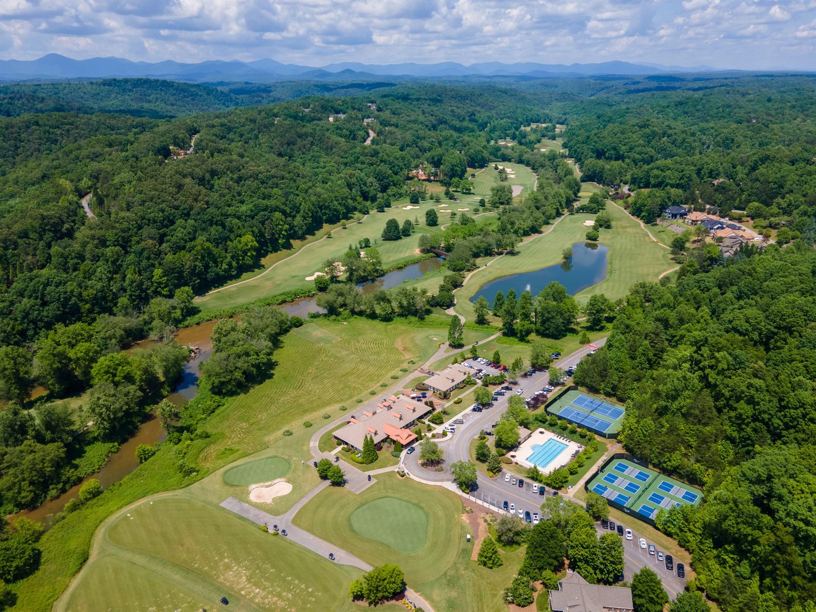 Aerial photograph of the Achasta amenity core in Dahlonega Georgia &mdash; clubhouse, pool, racquet courts, and surrounding fairways framed by mature hardwood canopy and the Chestatee River corridor