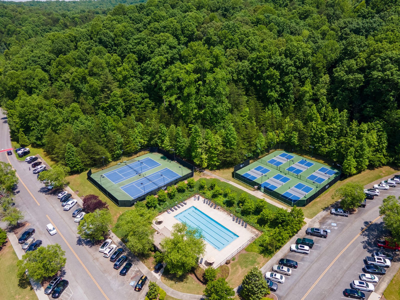 Top-down aerial of the Achasta amenity cluster showing the resort-style swimming pool, surrounding deck, tennis and pickleball courts, and adjacent firepit lounge inside the Achasta gates in Dahlonega Georgia
