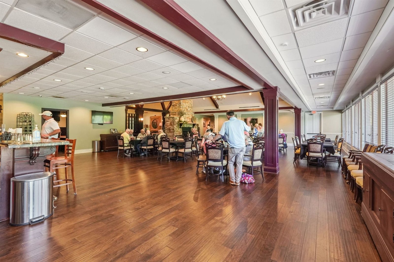 The Achasta Clubhouse dining hall &mdash; long, table-set dining room with upholstered chairs, hardwood floors, and tall arched windows looking onto the golf course &mdash; home of the Achasta Grill member dining program