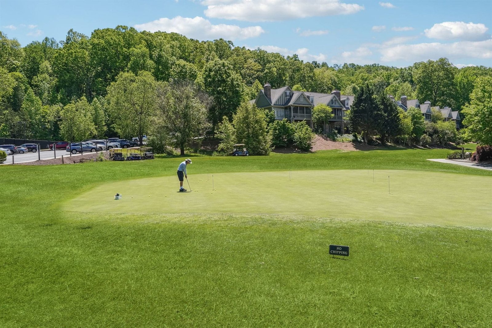 Large gabled stone and shake estate home behind the Achasta putting green &mdash; copper roof, multiple dormers, mature hardwood canopy surrounding the home, golf carts parked at left &mdash; representative of the residential character inside the gates