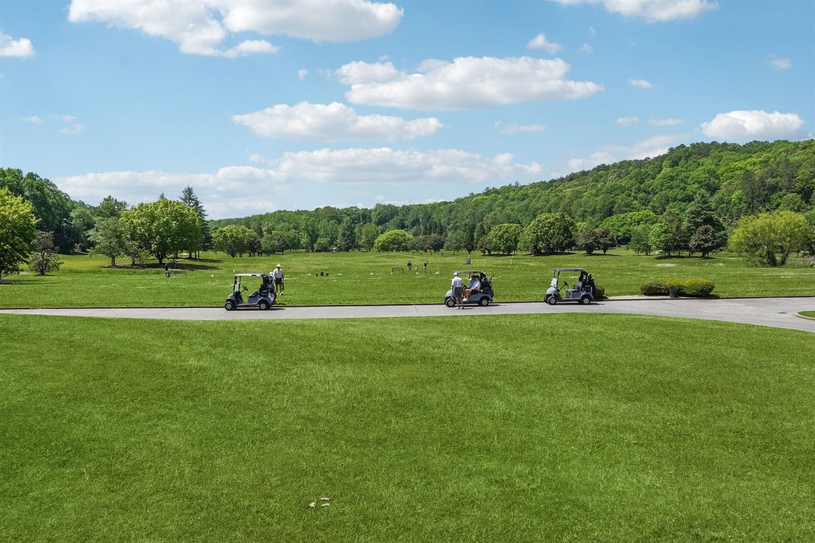 Four golf carts traveling along a paved cart path that crosses a fairway at Achasta &mdash; the back-nine routing between Chestatee River footbridges, with hardwood canopy framing the corridor on both sides