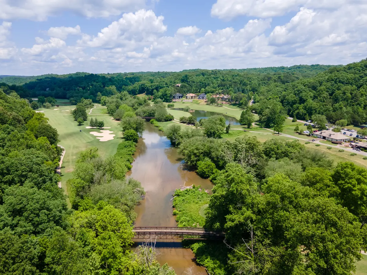 Aerial view of a river winding between dense green trees through the Achasta golf community, with a wooden footbridge crossing the river in the foreground, golf fairways and sand bunkers on the left, a pond, clubhouse with parking, and large homes on a wooded hillside beyond under a partly cloudy sky.