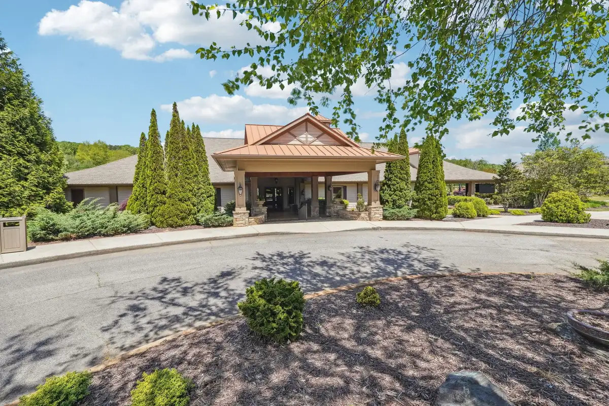 Front entrance of the Achasta community clubhouse on a sunny day: a copper metal-roofed portico with stone-based square columns over a covered entry, flanked by tall conifers and shrub landscaping, with a curving asphalt circular drive and mulched island in the foreground.