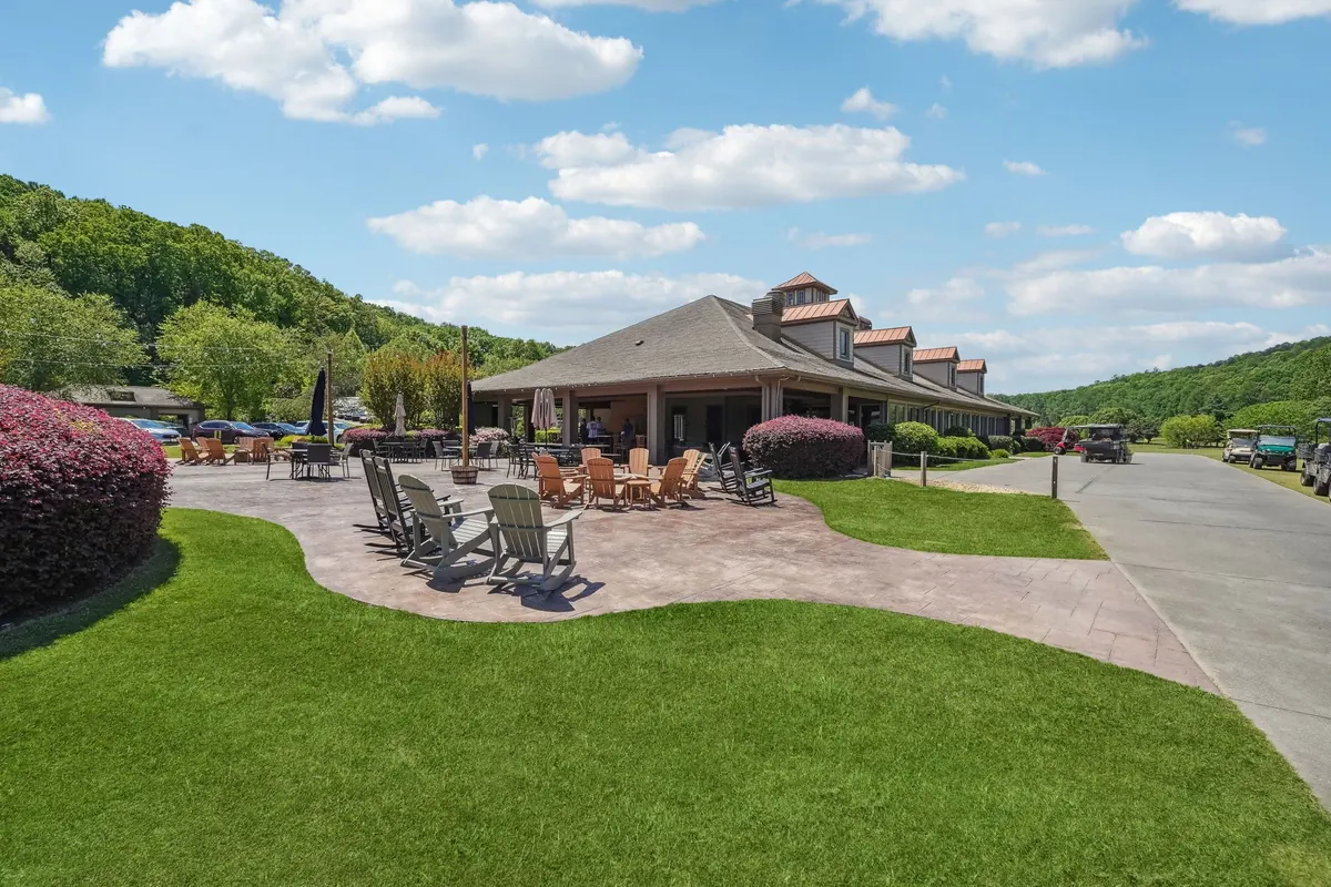 Clubhouse exterior from the cart-path side: stamped-concrete patio with Adirondack seating, low landscaping, paved cart path, hardwood-covered hills in the background.