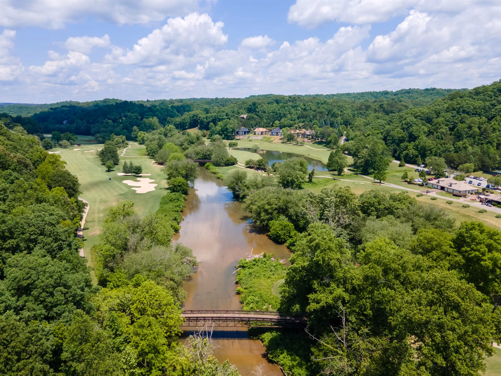Aerial view of the Chestatee River winding through the Achasta golf course with a pedestrian footbridge crossing the river and estate homes nestled in the hardwood canopy